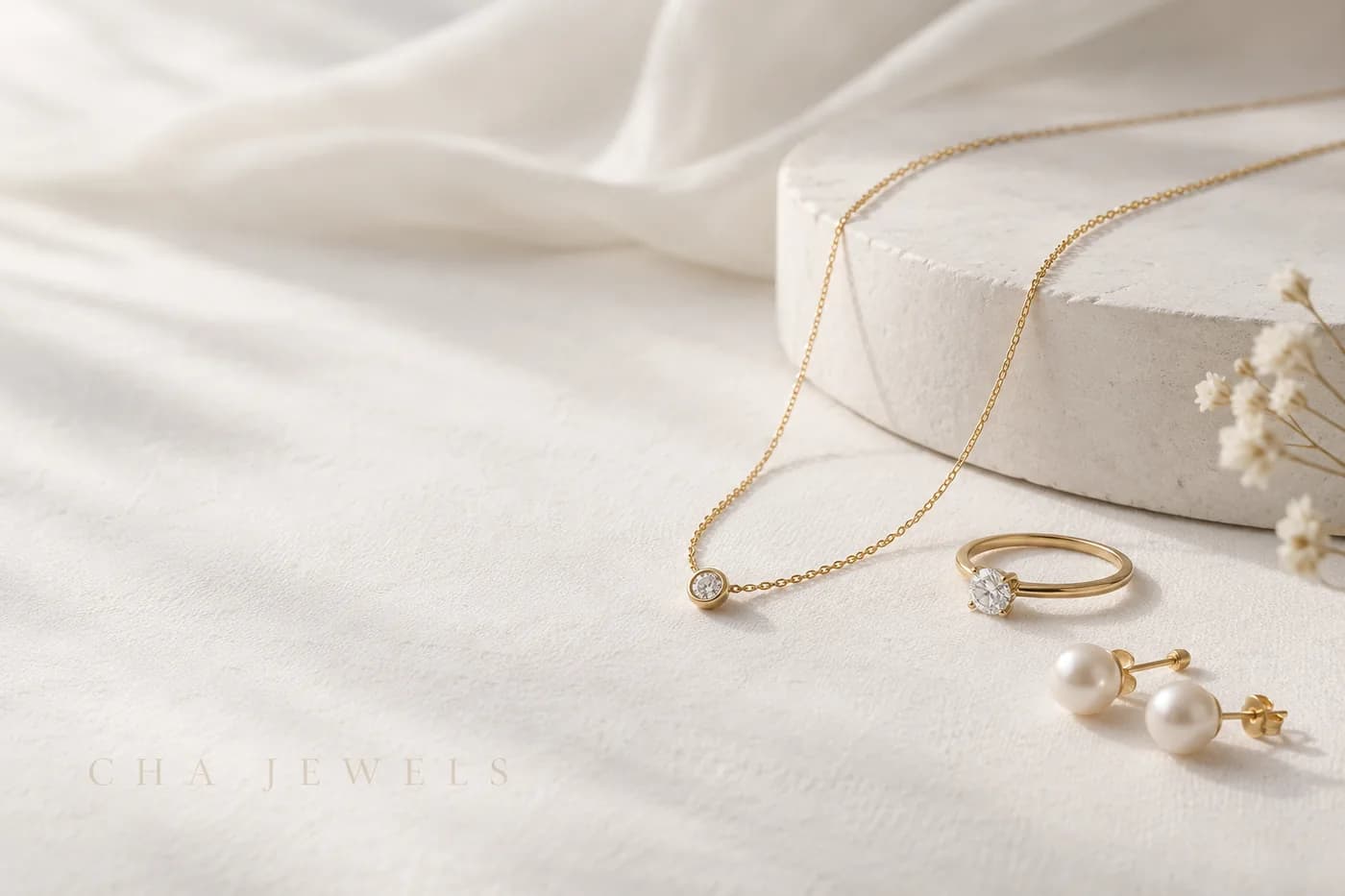 クリーム色の布の上に並べたCha Jewelsのゴールドネックレス、リング、パールピアス